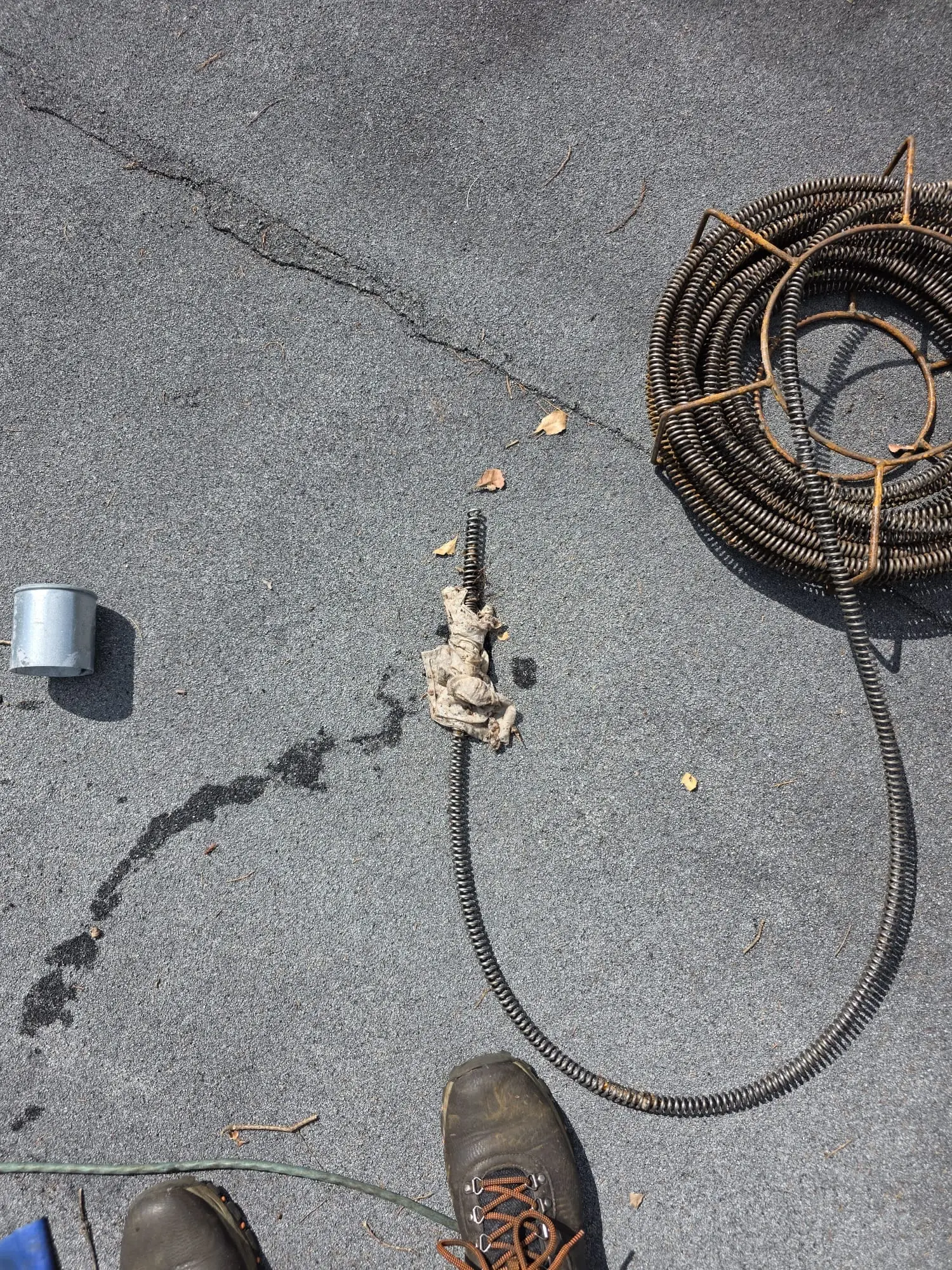 Coiled drain cable in a metal cage and an extended length of cable on asphalt, with a wad of cloth caught on its end and a metal pipe cap beside it