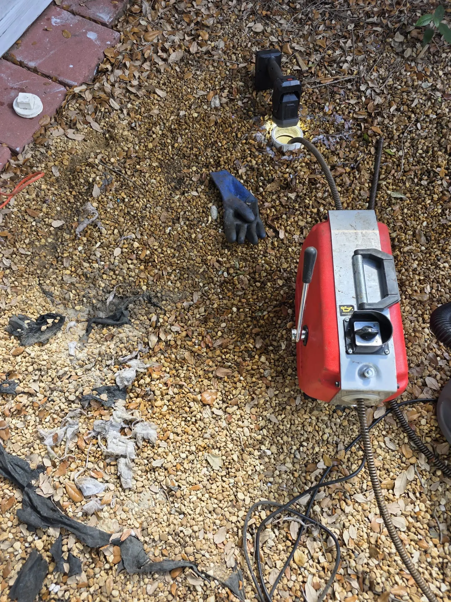 Red drain cable machine on a bed of small gravel with a cable running to an open white pipe, a blue work glove and wads of paper and cloth debris lying on the gravel, brick edging at the corner