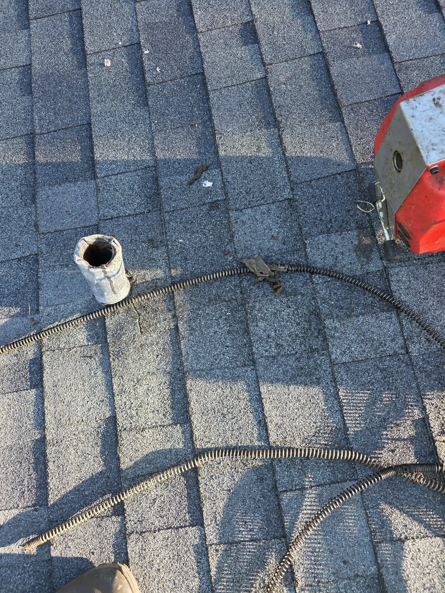 Drain cable fed through a vent pipe on a shingle roof, with a red drain machine at the edge of the frame and a dark wad of debris caught on the cable